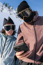 Two people in 686 Mens Gateway Shell Jackets, goggles, and beanies stand close together on a snowy mountain, looking at a smartphone with a bright blue sky behind them.