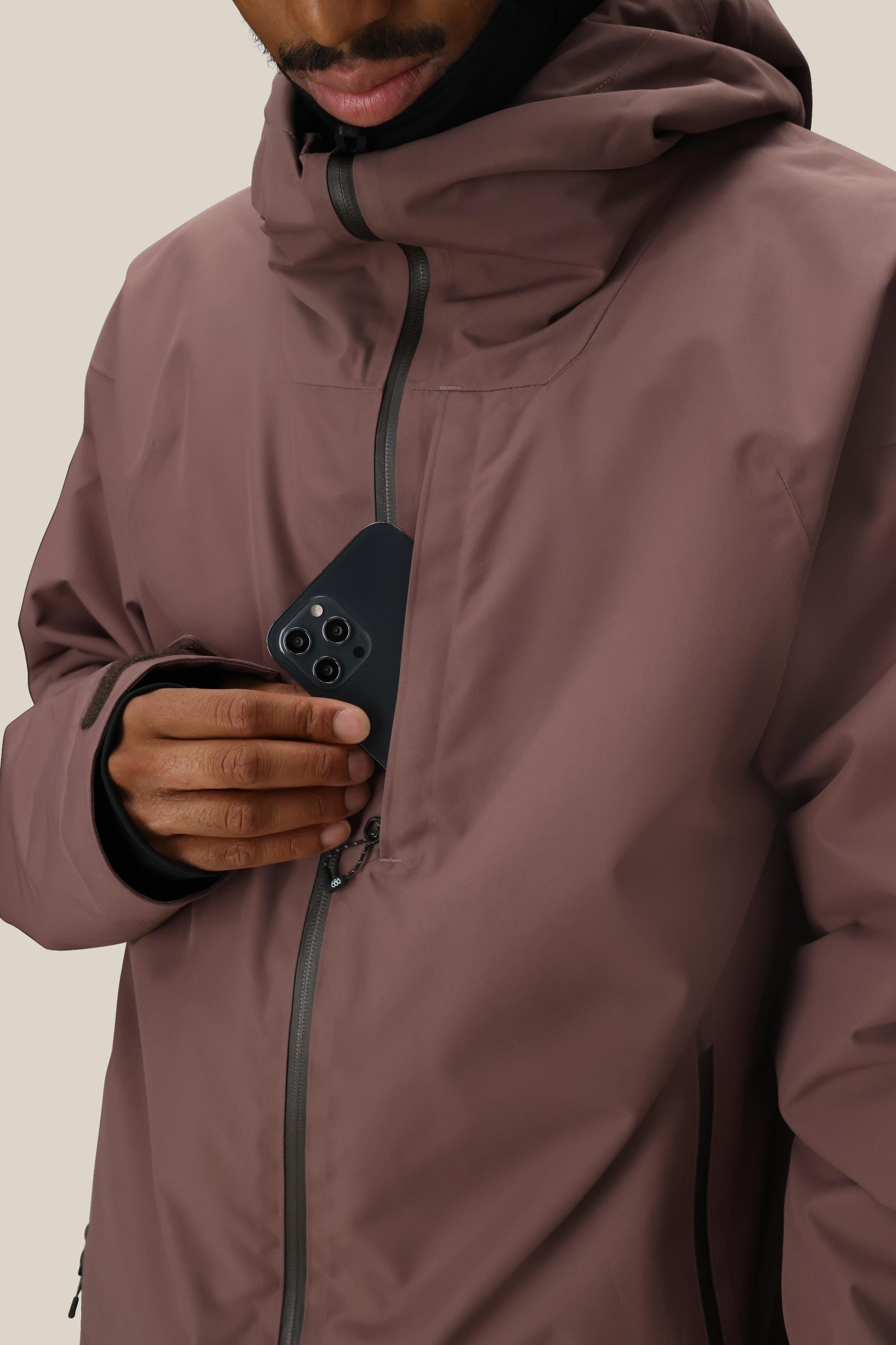 A man in a mauve 686 Mens Gateway Shell Jacket with a hood unzips the inner chest pocket and puts a black smartphone inside. His face is partially visible.
