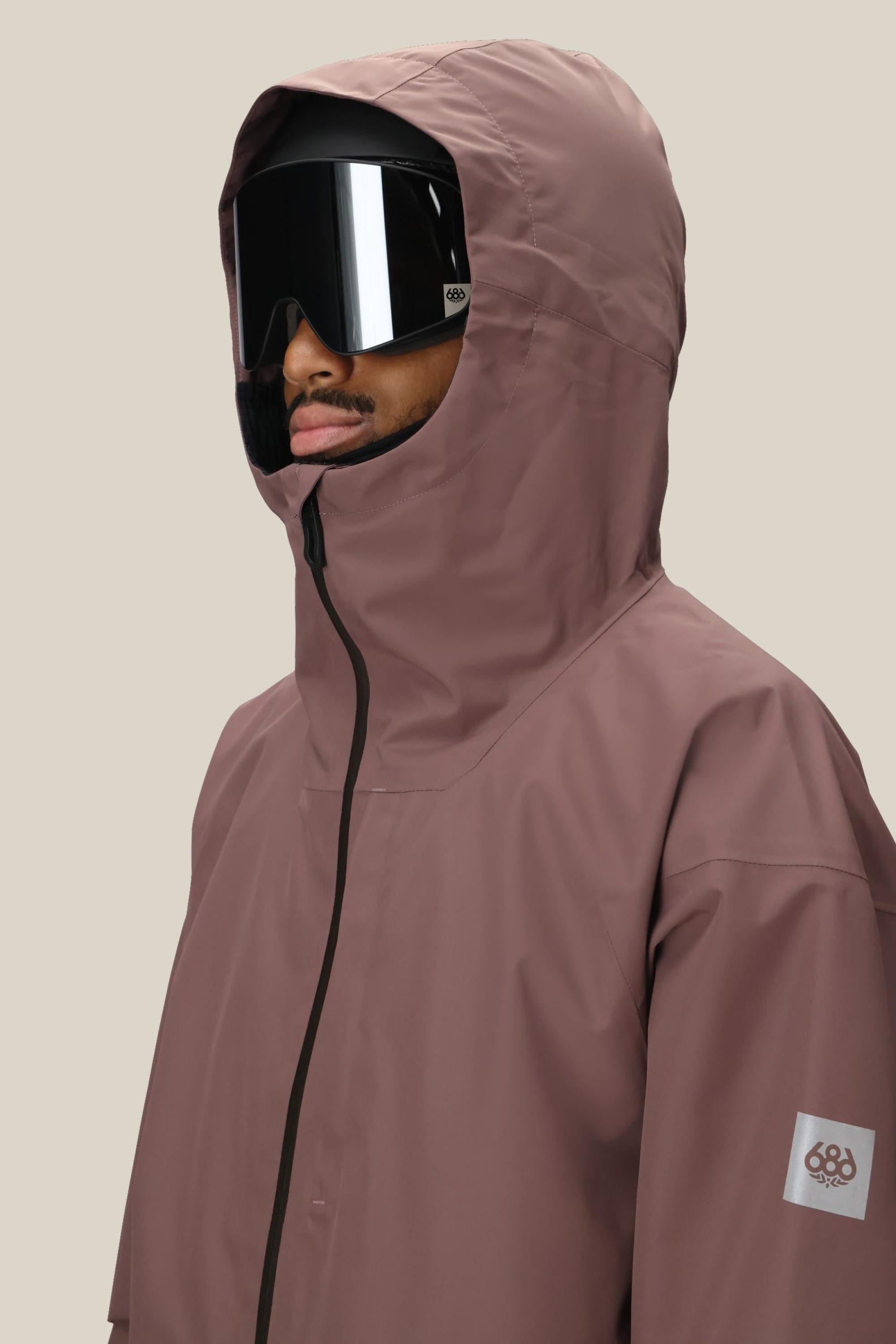 A person wears the 686 Mens Gateway Shell Jacket in mauve, featuring a 686 logo patch on the sleeve and zipper, paired with large black ski goggles. The hooded waterproof polyester jacket stands out against a plain beige background.