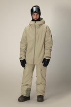 A man faces forward wearing the 686 Mens Gateway Shell Jacket by 686 in beige with a hood, matching pants, black gloves, dark boots, a black helmet, and ski goggles against a plain light background.