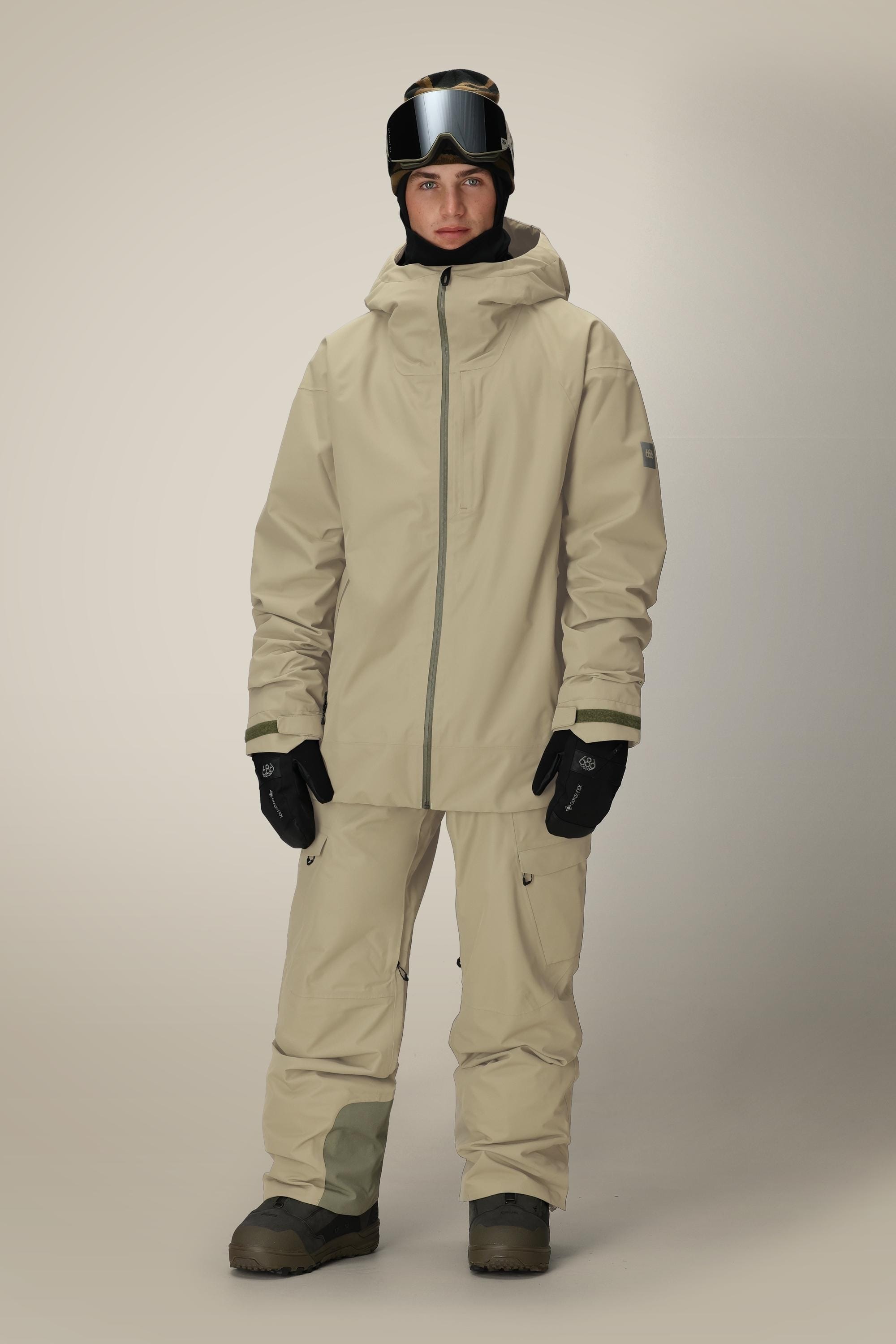 A man faces forward wearing the 686 Mens Gateway Shell Jacket by 686 in beige with a hood, matching pants, black gloves, dark boots, a black helmet, and ski goggles against a plain light background.