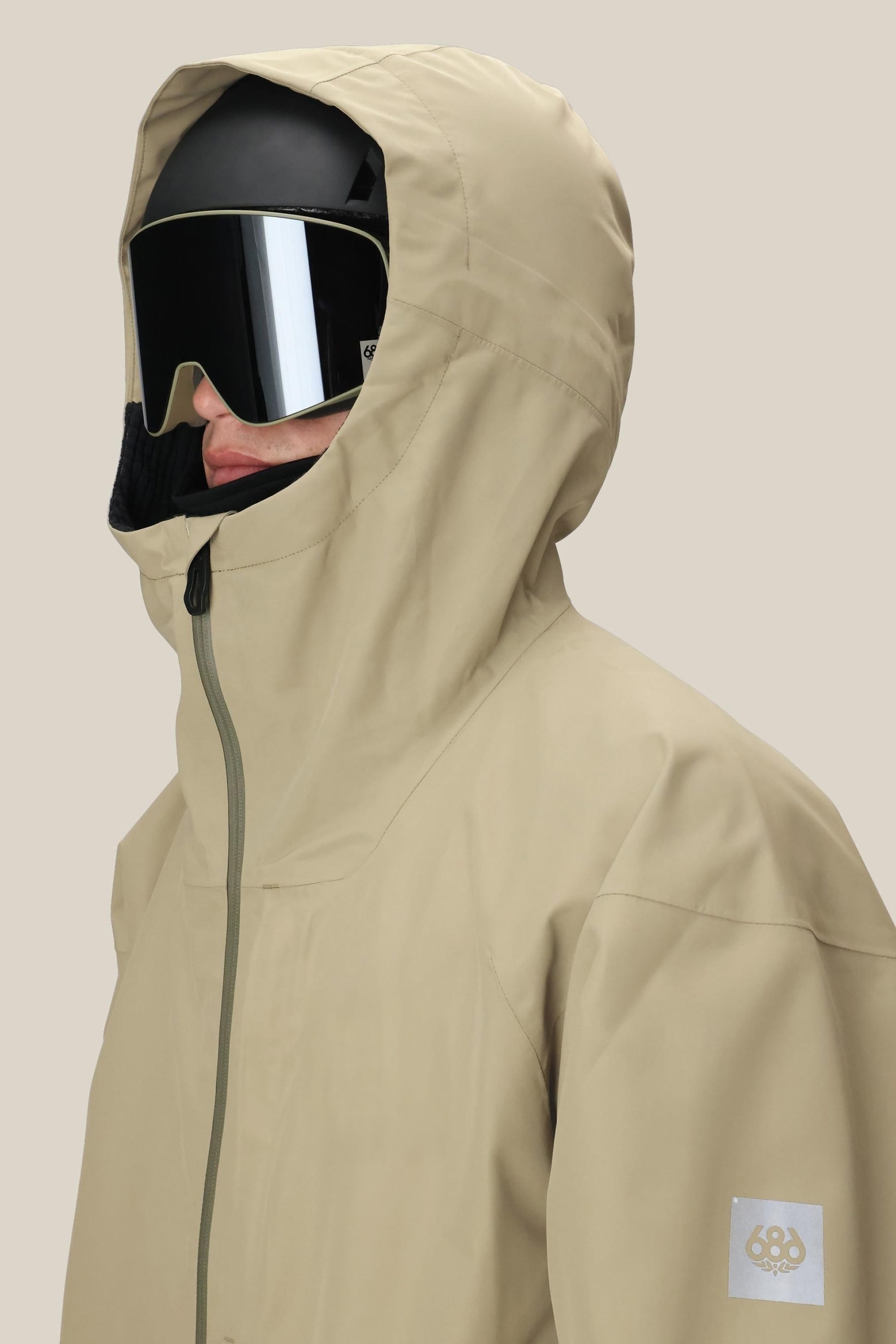 A person wearing the 686 Mens Gateway Shell Jacket in beige with a high collar, paired with a black ski helmet and large reflective ski goggles, faces left against a plain light background.