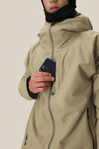 A person in a beige 686 Mens Gateway Shell Jacket with a hood and black face covering puts a black smartphone into the jacket’s chest pocket, set against a neutral background.