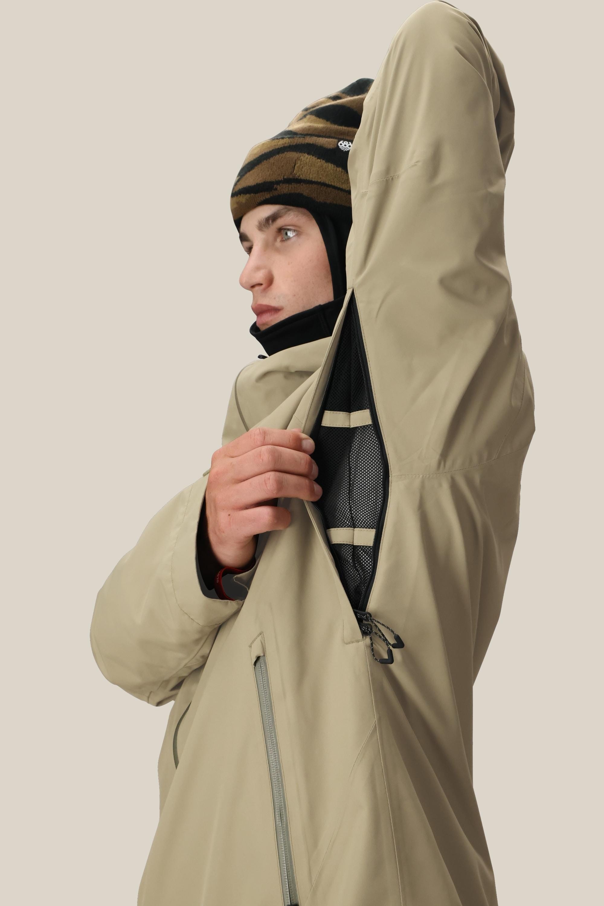 A man in a 686 Mens Gateway Shell Jacket and a striped beanie unzips the mesh-lined underarm vent, showcasing the jackets ventilation feature against a plain, light background.