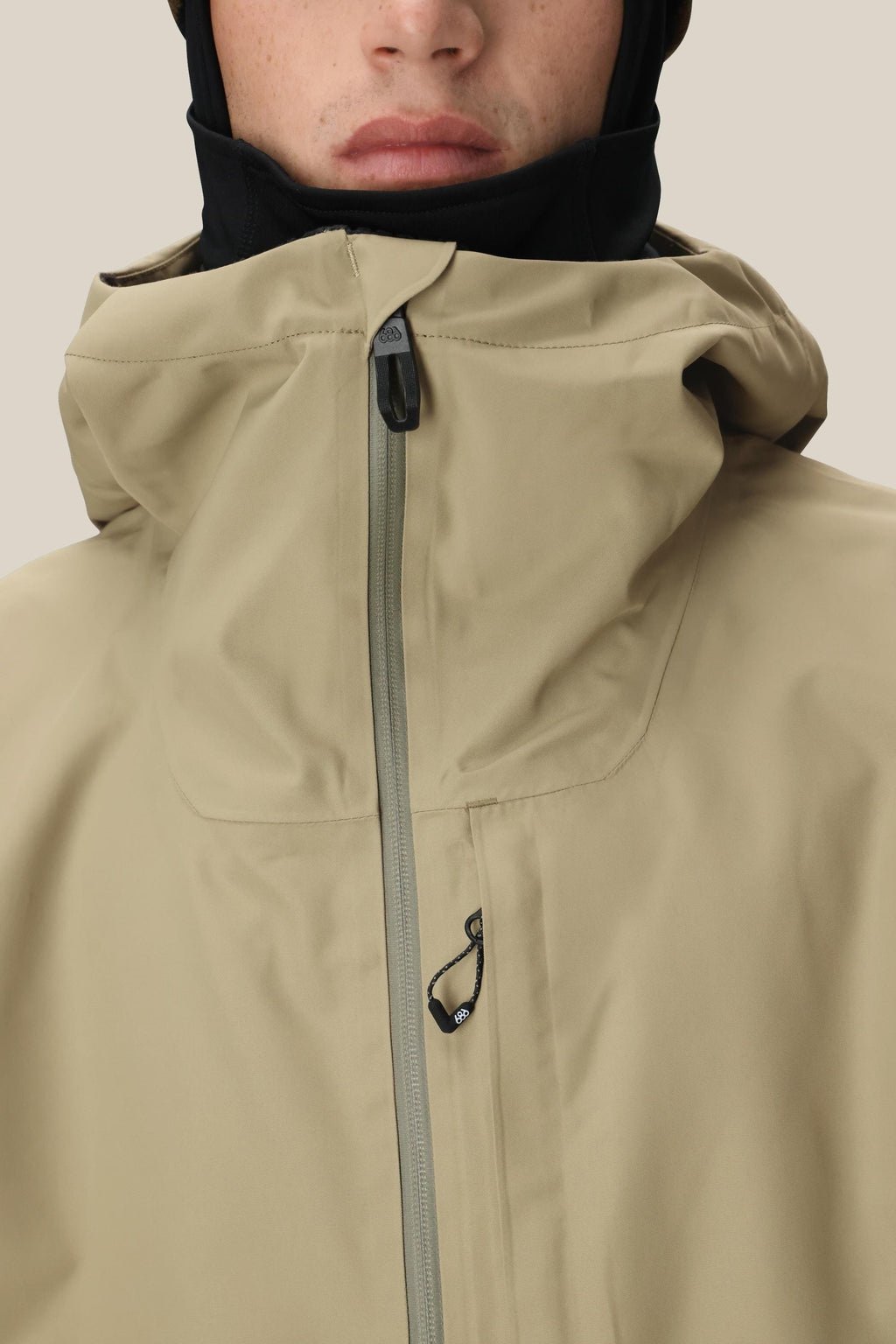 A close-up of a person wearing the 686 Mens Gateway Shell Jacket in beige, featuring a high collar and front zipper that partially covers their face. A black underlayer covers their neck and head, showcasing this functional waterproof jacket from 686.