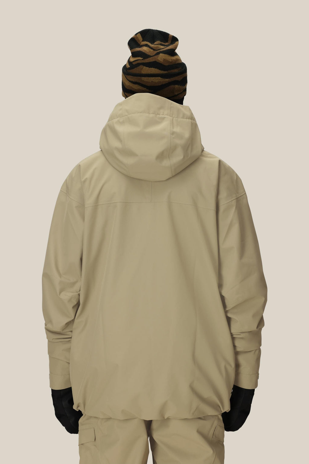 A person faces away, wearing the 686 Mens Gateway Shell Jacket in beige by 686 with a hood, matching pants, black gloves, and a brown-black striped beanie against a plain light background.