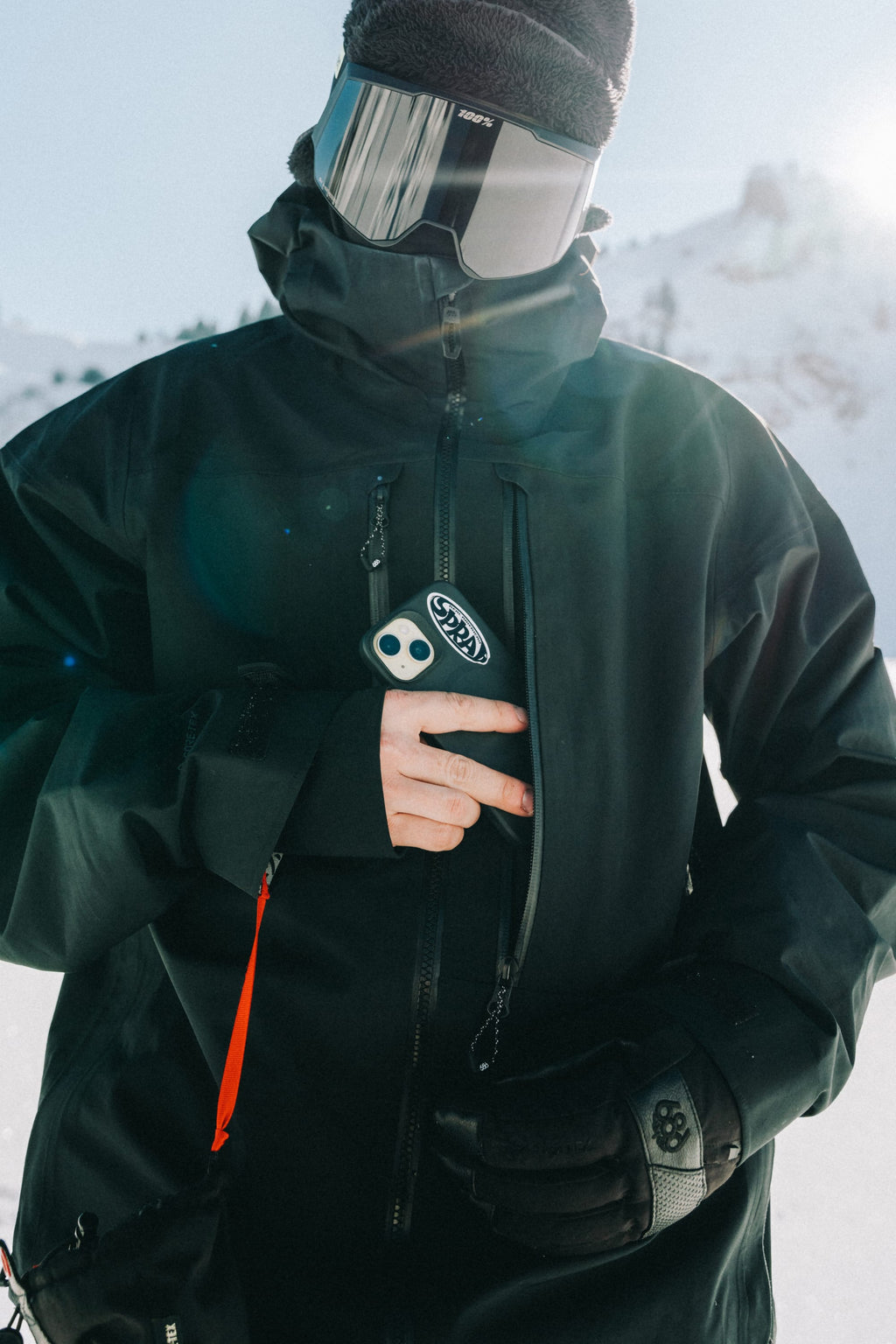 A person wearing the 686 Men's GORE-TEX 3L ATV Thermagraph® Jacket and reflective goggles holds an avalanche beacon while standing in a snowy mountain landscape.