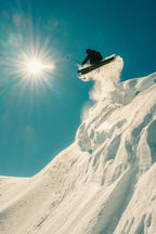 A skier wearing the 686 Men's GORE-TEX 3L ATV Thermagraph® Jacket soars off a snowy cliff under a clear blue sky, sunlight in the background and powdery snow trailing behind.
