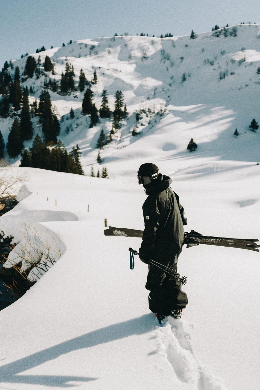 Wearing the 686 Men's GORE-TEX 3L ATV Thermagraph® Jacket, a person with a helmet carries skis while trekking through deep snow in a snowy mountain landscape dotted with pine trees.