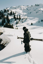 Wearing the 686 Men's GORE-TEX 3L ATV Thermagraph® Jacket, a person with a helmet carries skis while trekking through deep snow in a snowy mountain landscape dotted with pine trees.