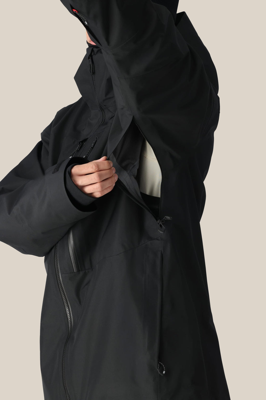 A man in a 686 Men's GORE-TEX 3L ATV Thermagraph® Jacket unzips an underarm vent, showing part of a light inner layer. The black polyester jacket by 686 features multiple zippers and a high collar.