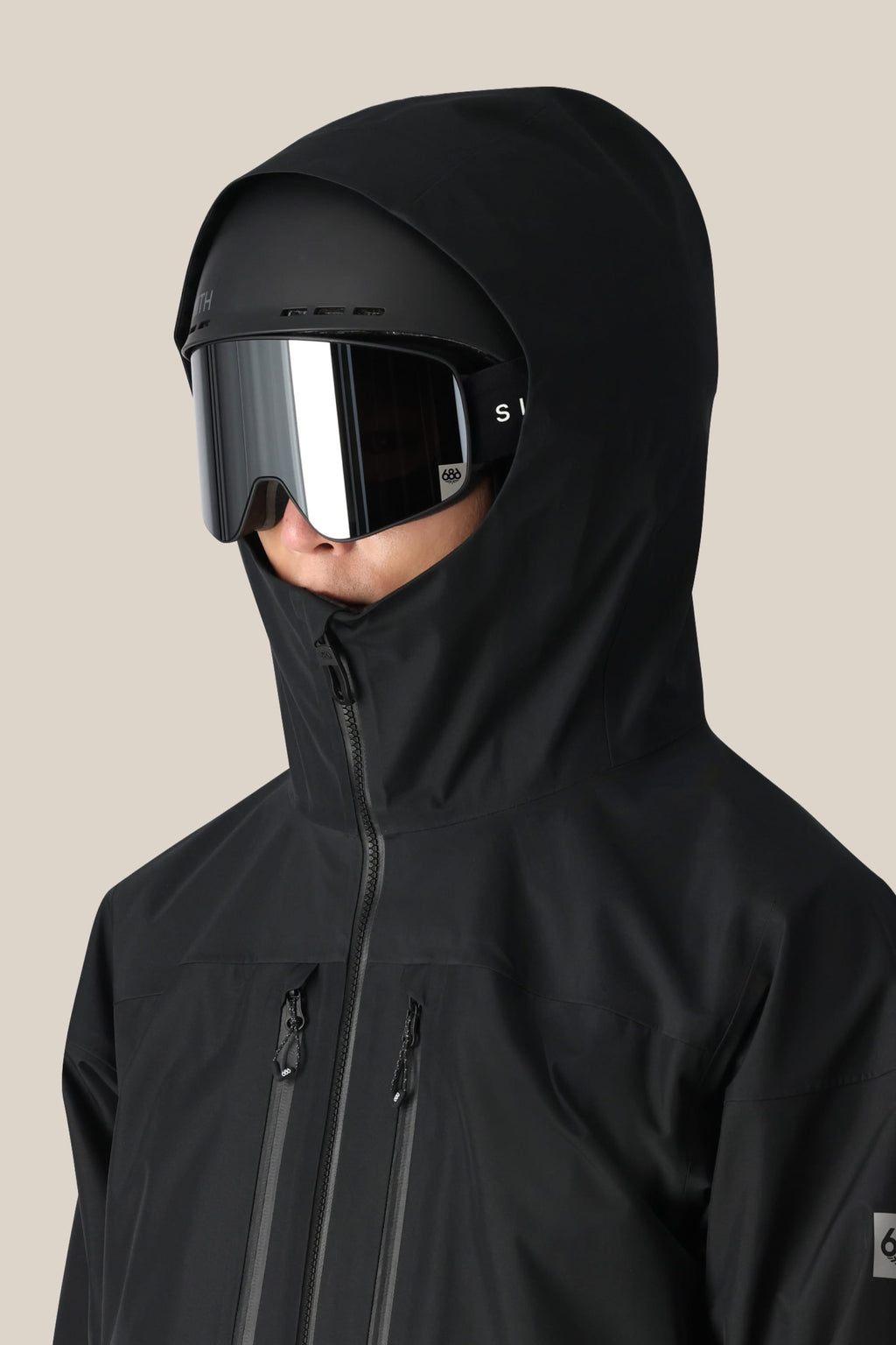 A person wearing the 686 Men's GORE-TEX 3L ATV Thermagraph® Jacket with the hood up and large, reflective ski goggles faces slightly right against a plain beige background.