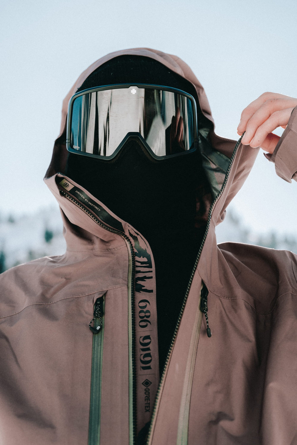 A person outdoors in snowy mountains partially unzips their brown 686 Men's GORE-TEX 3L ATV Thermagraph® Jacket, wearing a black ski mask and reflective ski goggles.