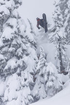 A snowboarder in the 686 Men's GORE-TEX 3L ATV Thermagraph® Jacket launches into a mid-air trick amid tall, snow-covered pines. Thick snow blankets the trees and ground, capturing a dramatic winter scene.