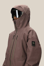 A person wears a 686 Men's GORE-TEX 3L ATV Thermagraph® Jacket in mauve, featuring zipped pockets and a GORE-TEX patch on the sleeve, paired with large black ski goggles. The background is plain beige.