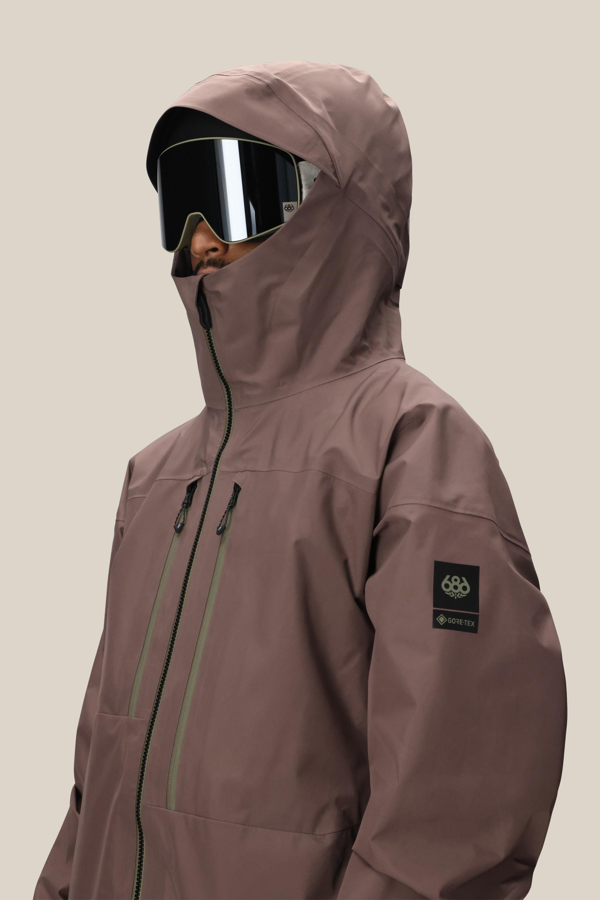 A person wears a 686 Men's GORE-TEX 3L ATV Thermagraph® Jacket in mauve, featuring zipped pockets and a GORE-TEX patch on the sleeve, paired with large black ski goggles. The background is plain beige.