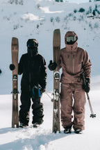Two people in ski gear and goggles stand in the snow, each holding skis and poles. One wears the 686 Men's GORE-TEX 3L ATV Thermagraph® Jacket by 686 as they pose with snow-covered mountains and trees behind them.