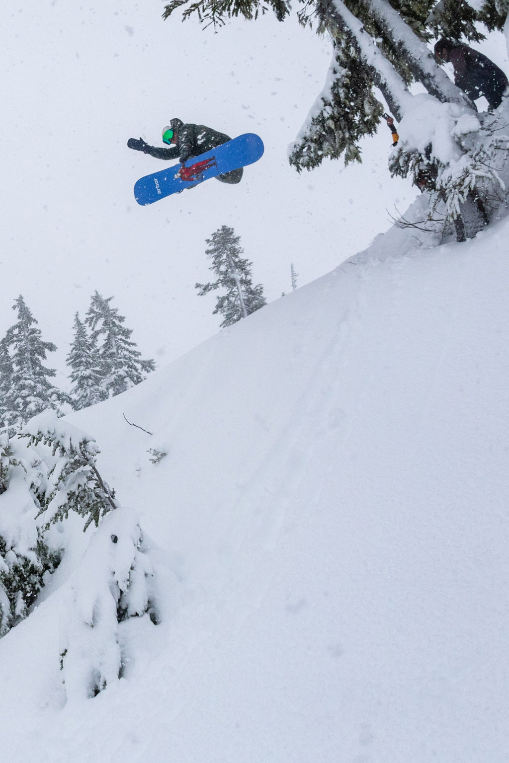 A snowboarder in a 686 Men's Nightline™ Insulated Jacket soars off a snowy slope, surrounded by snow-covered trees in a wintry landscape.