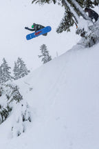 A snowboarder in a 686 Men's Nightline™ Insulated Jacket soars off a snowy slope, surrounded by snow-covered trees in a wintry landscape.