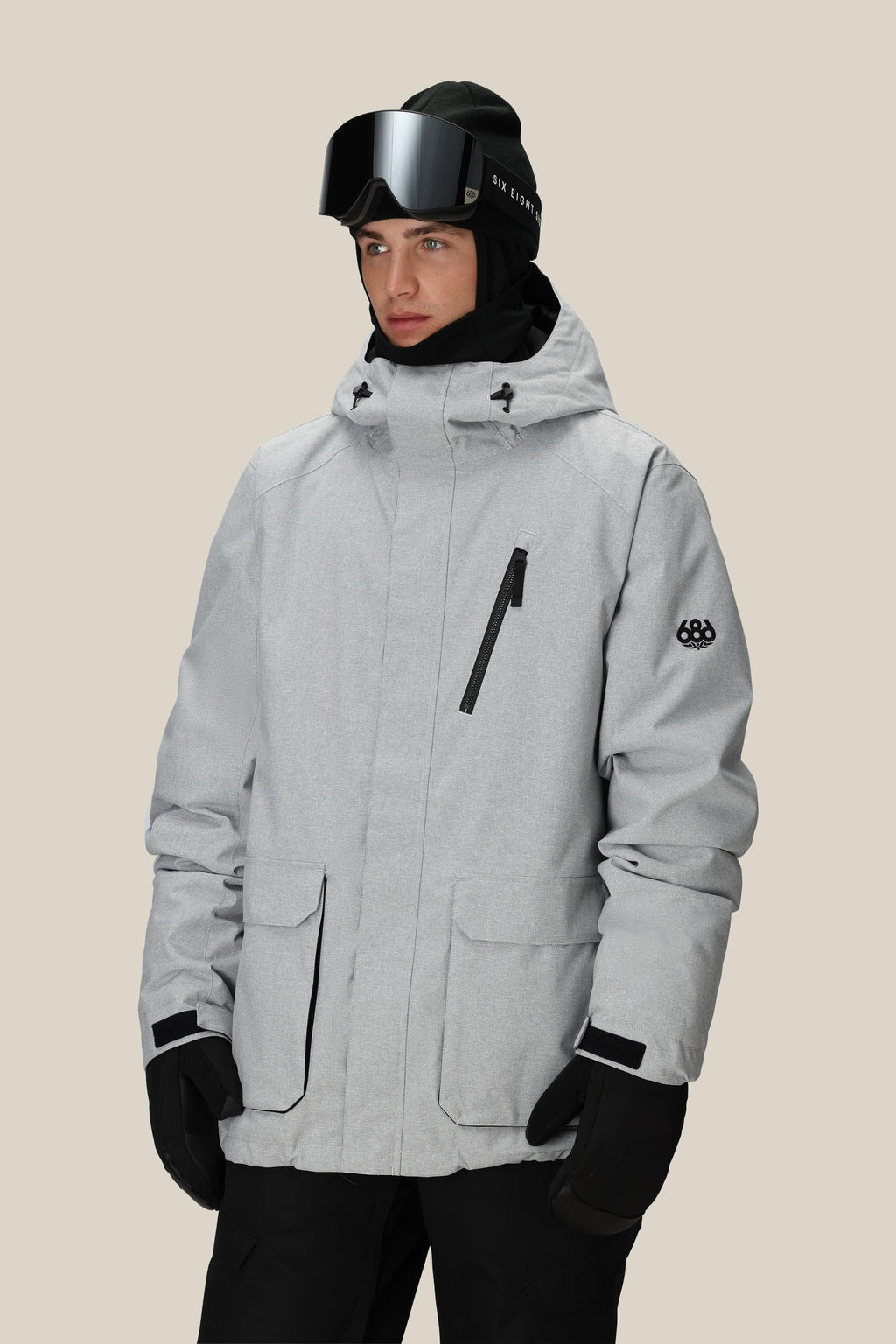 A person wearing the 686 Men's Nightline™ Insulated Jacket in light gray, paired with black gloves, black ski goggles, and a black hat, stands against a plain beige background.