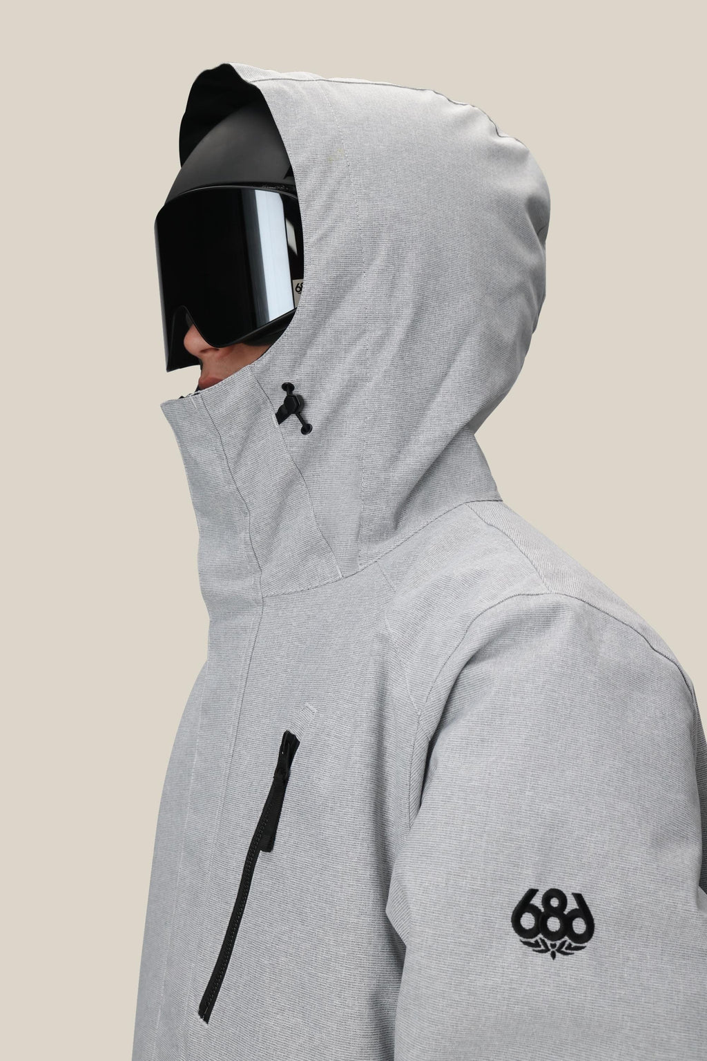 A person in profile wears the 686 Men's Nightline™ Insulated Jacket in light gray, featuring a black sleeve logo, chest zipper pocket, and large black ski goggles, set against a plain beige background.