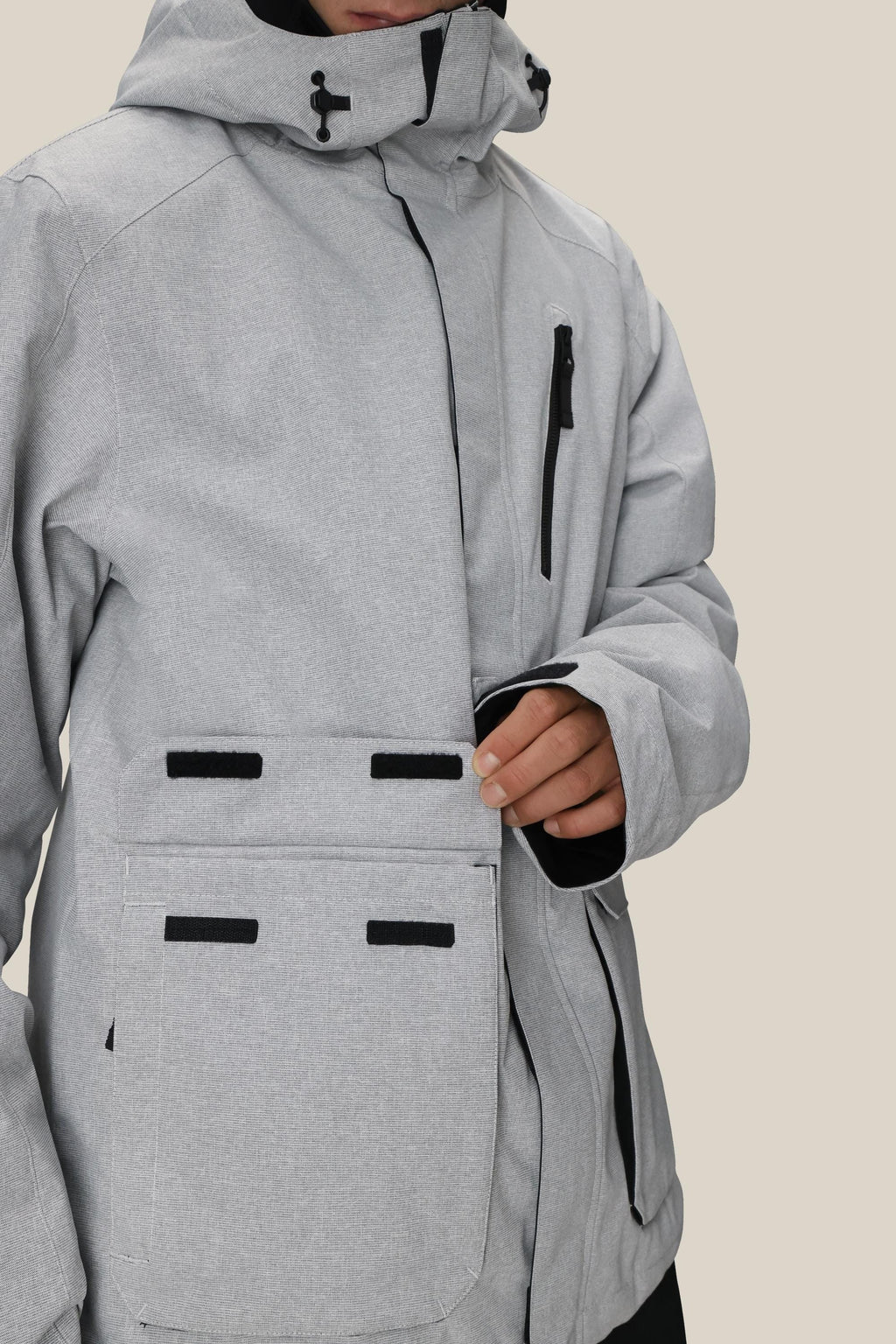 A man fastens the velcro flap on a large front pocket of the 686 Men's Nightline™ Insulated Jacket, featuring a light gray color with black zipper and velcro accents by 686.