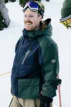 A man wearing the 686 Men's Nightline™ Insulated Jacket stands outdoors in the snow, smiling with ski goggles on his head and holding ski poles. Evergreen trees and a snowy landscape provide the backdrop.