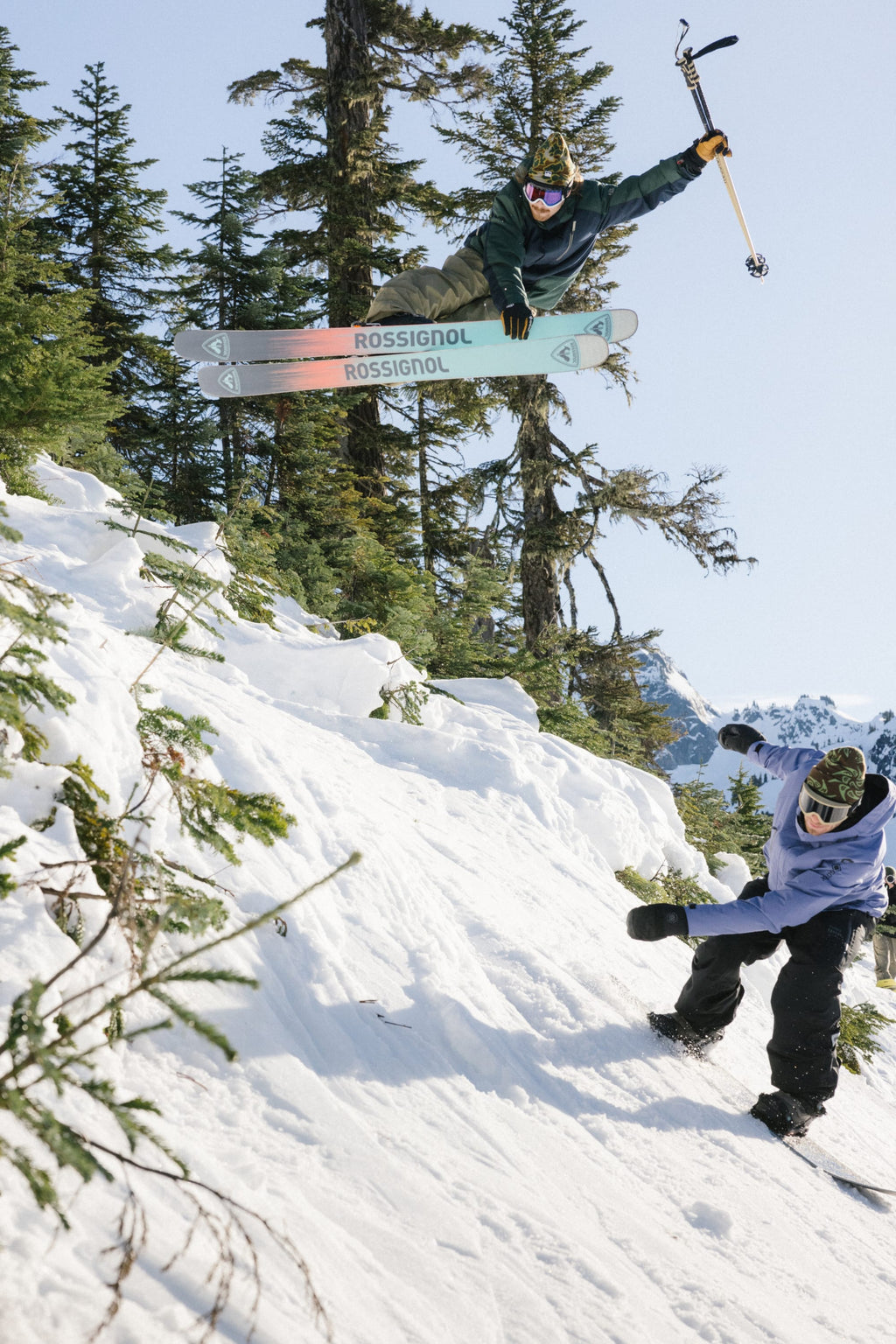 A skier in the 686 Men's Nightline™ Insulated Jacket jumps off a snowy slope with skis spread and an ice axe raised, as a snowboarder in a blue jacket rides downhill below. Evergreen trees and mountains fill the background.