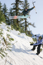 A skier in the 686 Men's Nightline™ Insulated Jacket jumps off a snowy slope with skis spread and an ice axe raised, as a snowboarder in a blue jacket rides downhill below. Evergreen trees and mountains fill the background.