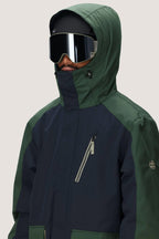 A person wears the 686 Men's Nightline™ Insulated Jacket in green and dark blue, paired with large reflective ski goggles and a black helmet. The hood is up and a plain light gray background highlights the stylish outerwear from 686.