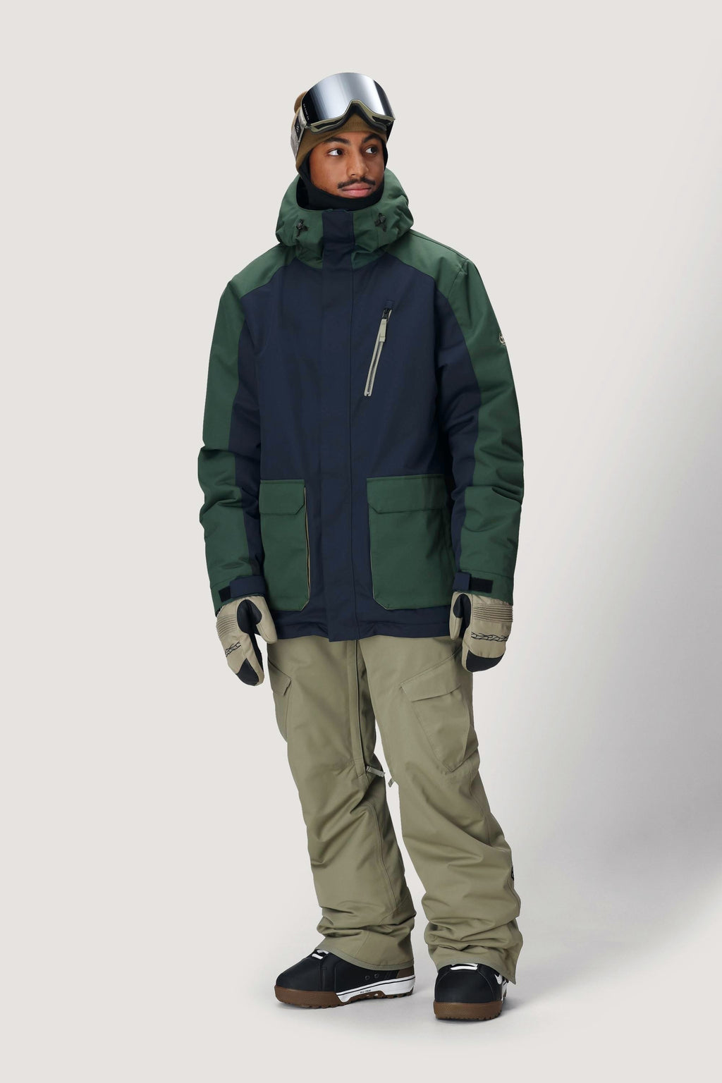 A man in the 686 Men's Nightline™ Insulated Jacket (navy and green) with khaki snow pants, gloves, boots, and ski goggles stands against a plain light background, perfectly outfitted for winter sports.
