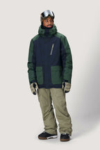 A man in the 686 Men's Nightline™ Insulated Jacket (navy and green) with khaki snow pants, gloves, boots, and ski goggles stands against a plain light background, perfectly outfitted for winter sports.