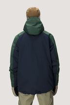 Wearing a brown beanie and the 686 Men's Nightline™ Insulated Jacket—featuring dark green sleeves, navy blue back, and hood—with beige pants, a person stands facing away from the camera against a light gray background.