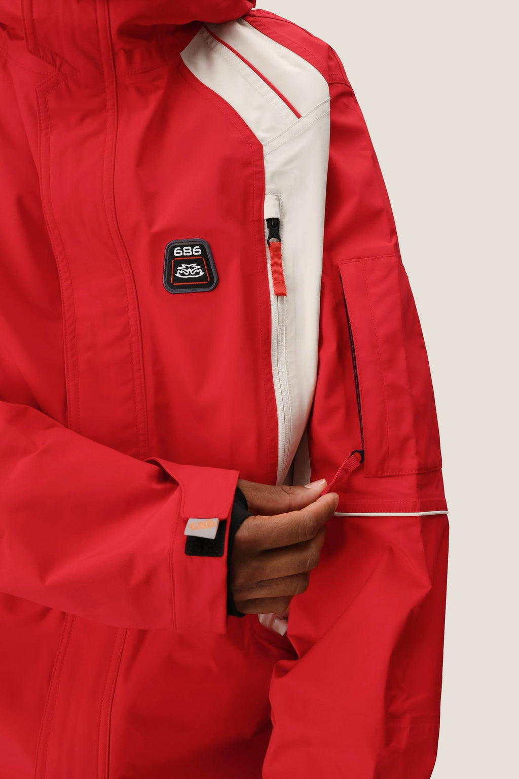 A person wearing the 686 Men's 2002 X3 3L Shell Jacket unzips a sleeve pocket. The red and white waterproof jacket features a chest “686” patch and Velcro strap cuffs.