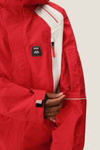 A person wearing the 686 Men's 2002 X3 3L Shell Jacket unzips a sleeve pocket. The red and white waterproof jacket features a chest “686” patch and Velcro strap cuffs.