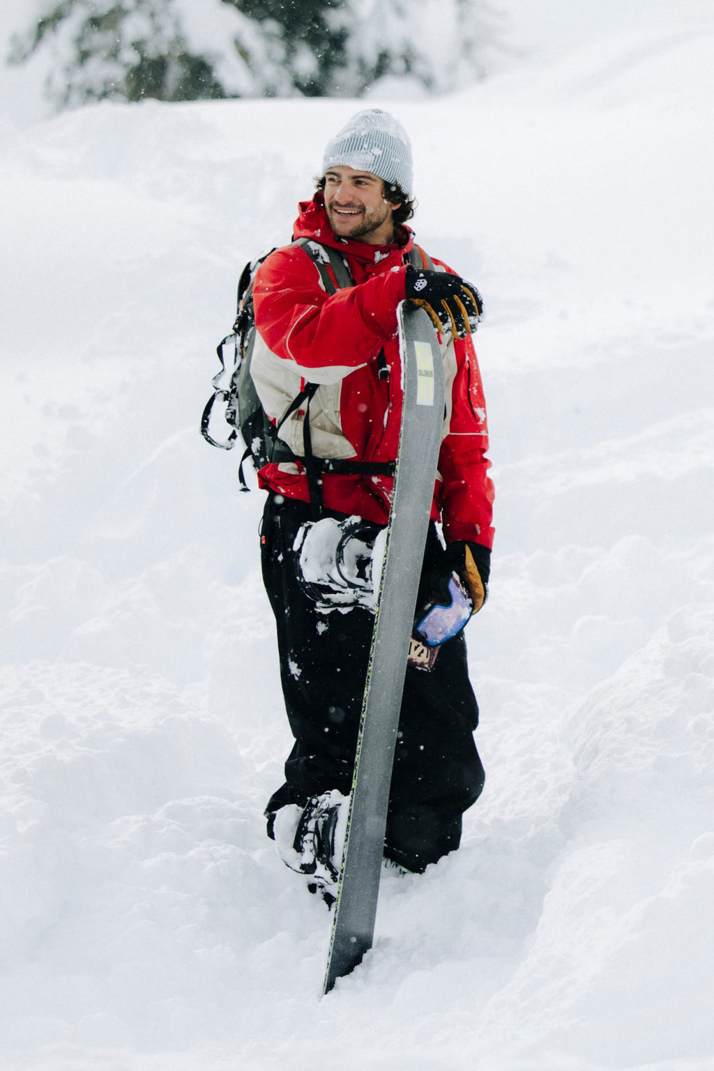 A person in a 686 Men's 2002 X3 3L Shell Jacket and gray beanie stands smiling in deep snow, holding a snowboard upright with snowy trees in the background.