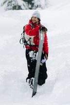A person in a 686 Men's 2002 X3 3L Shell Jacket and gray beanie stands smiling in deep snow, holding a snowboard upright with snowy trees in the background.