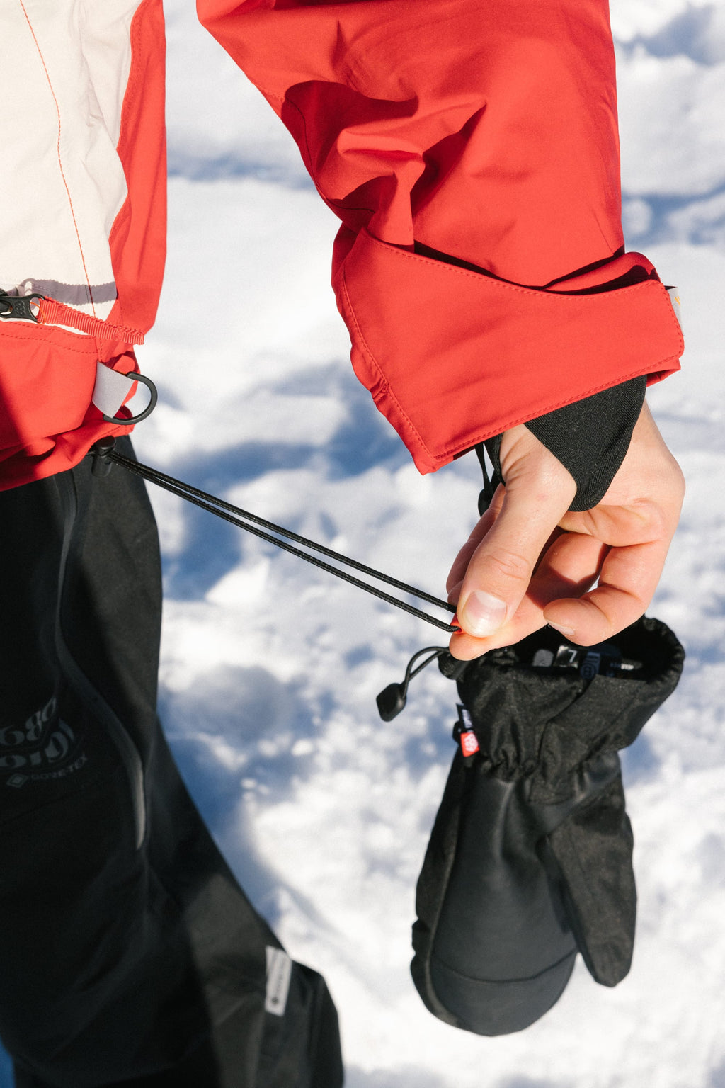 Wearing the 686 Men's 2002 X3 3L Shell Jacket, a person adjusts a black drawstring on their winter pants and holds black gloves, with snow in the background.