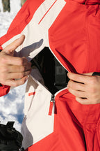 A person in a 686 Mens 2002 X3 Shell Jacket unzips a pocket, revealing a black strap inside while snow falls in the background.