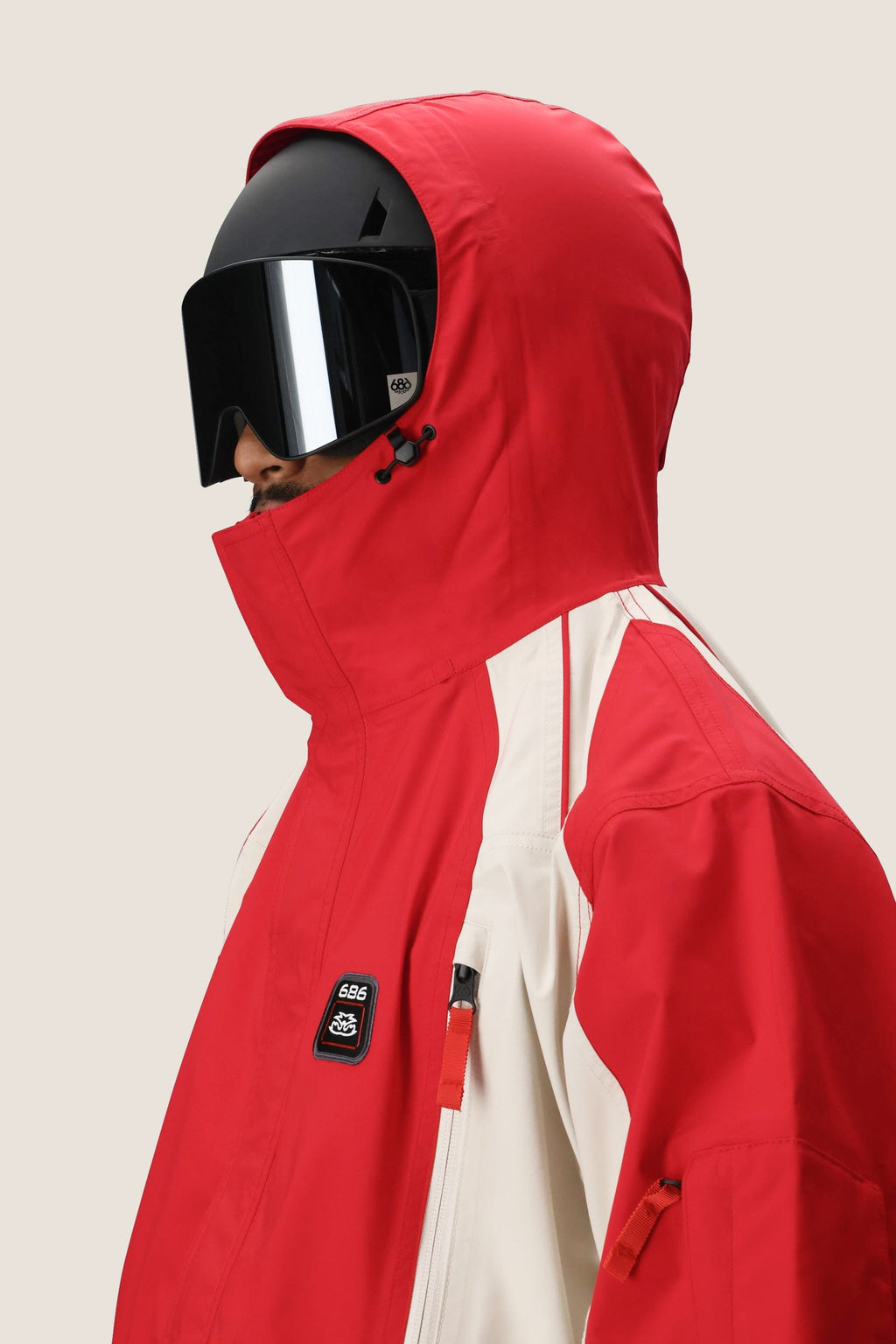 A person wearing the 686 Men's 2002 X3 3L Shell Jacket in red and white by 686, featuring a high collar, paired with large black ski goggles, faces left against a plain light background.