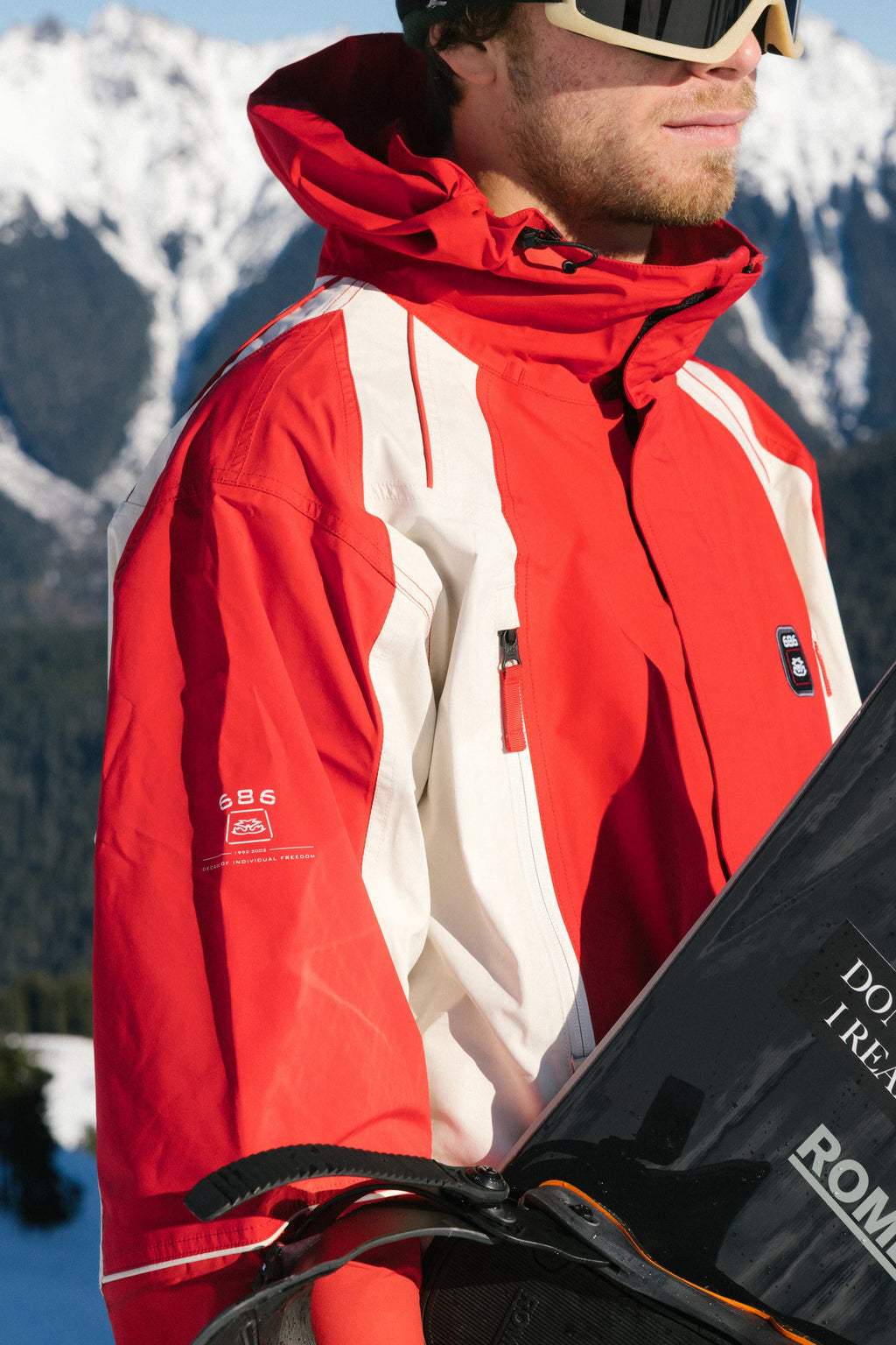 A person in a 686 Men's 2002 X3 3L Shell Jacket in red and white with ski goggles holds a snowboard, standing outdoors before snowy mountains.