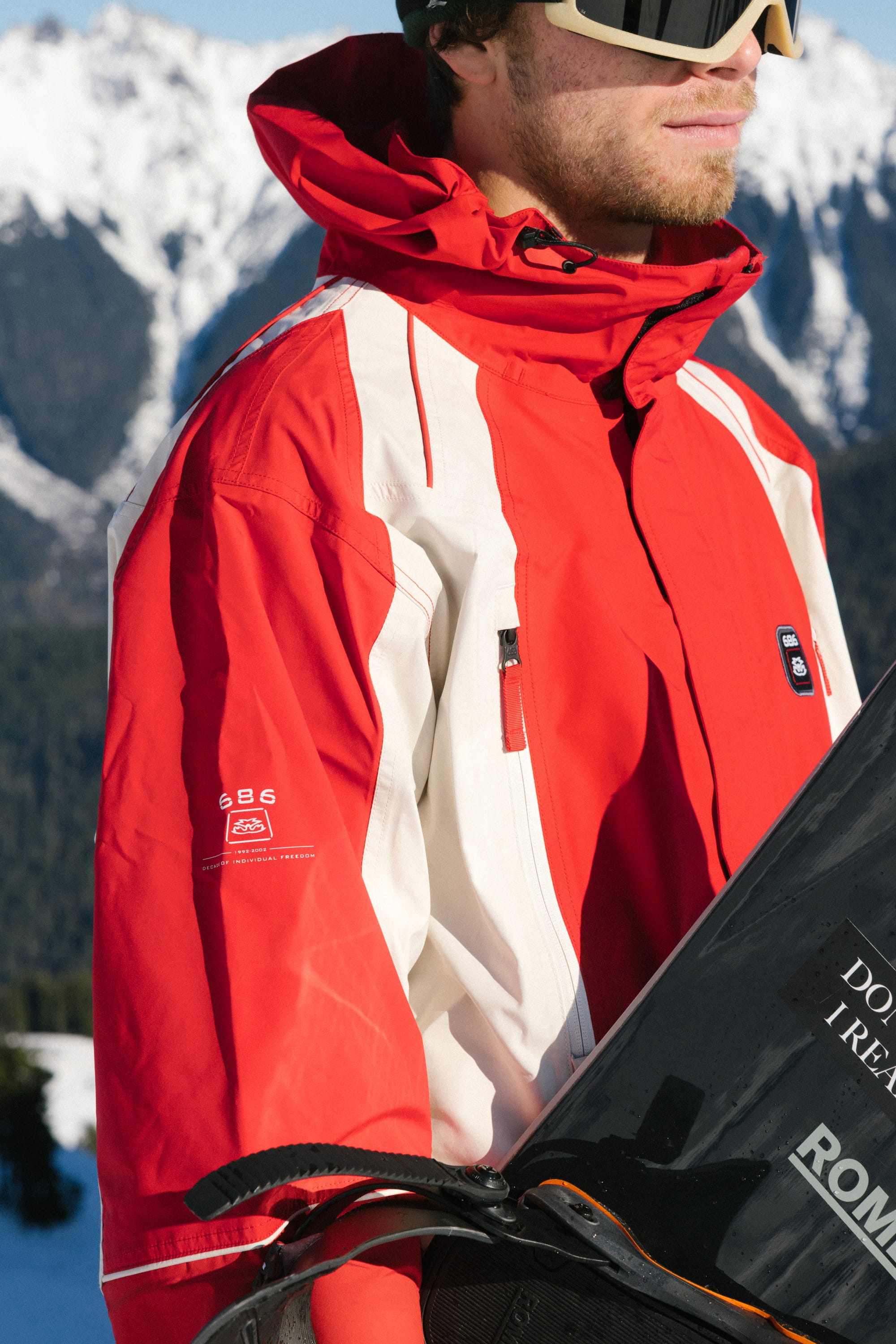 A person in a 686 Men's 2002 X3 3L Shell Jacket in red and white with ski goggles holds a snowboard, standing outdoors before snowy mountains.