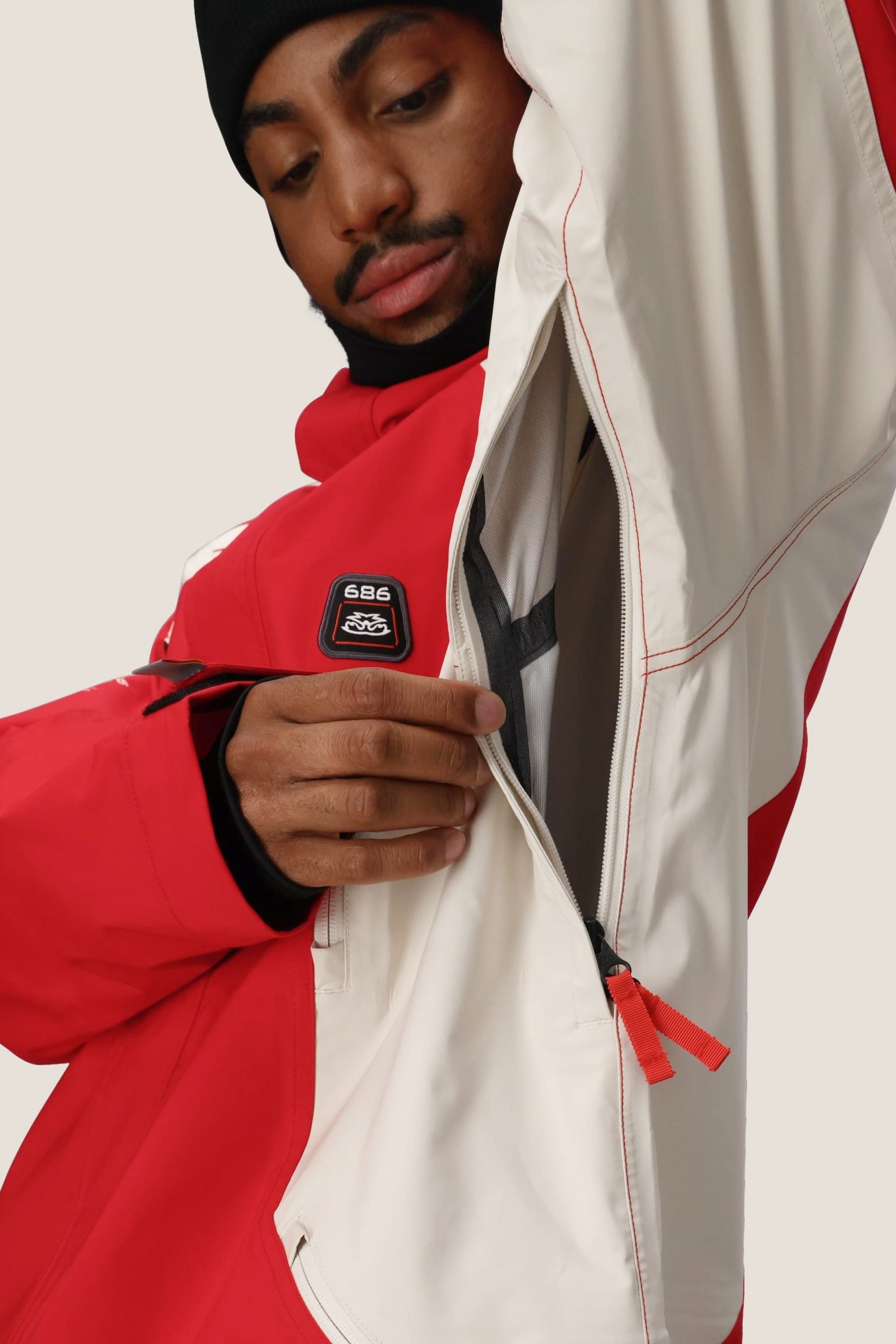 A person in a red and white 686 Men's 2002 X3 3L Shell Jacket unzips the large side vent under the arm, revealing part of the lining. They wear a black hat and look down at the zipper.