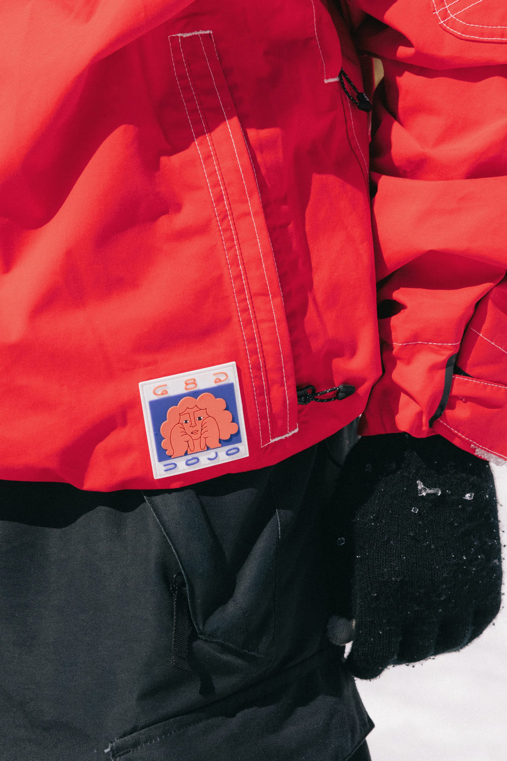 A person stands outdoors in the snow wearing the 686 Dojo® GORE-TEX Jacket in red with a colorful brain patch, paired with black pants and a black glove.