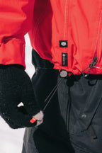 A person wearing the 686 Dojo® GORE-TEX Jacket by 686 and black gloves tightens the jacket's drawstring at the waist while standing outdoors in a snowy setting.