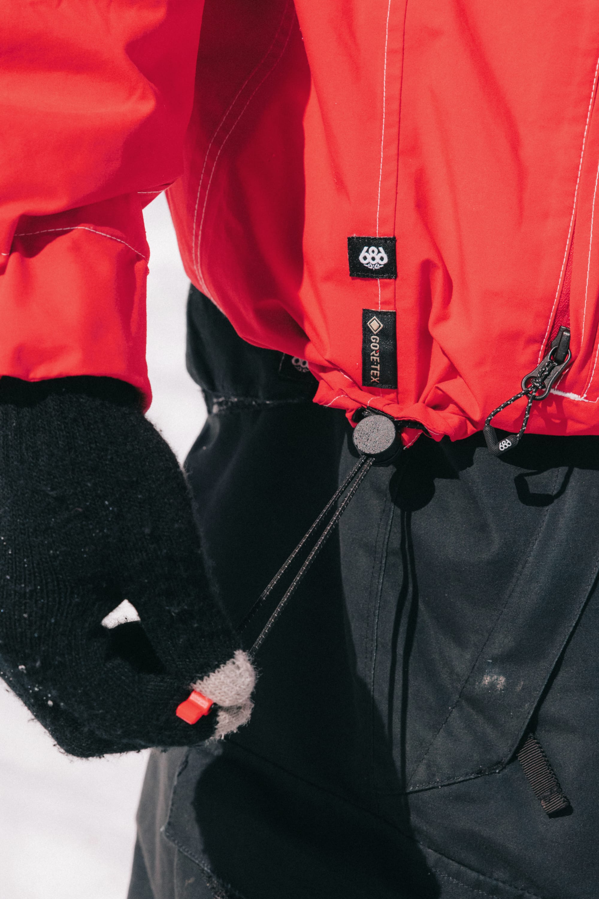 A person wearing the 686 Dojo® GORE-TEX Jacket by 686 and black gloves tightens the jacket's drawstring at the waist while standing outdoors in a snowy setting.