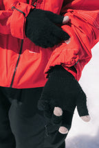 A person wearing the 686 Dojo® GORE-TEX Jacket and red and black touchscreen gloves with gray fingertips stands on snow, adjusting one glove with the other hand.