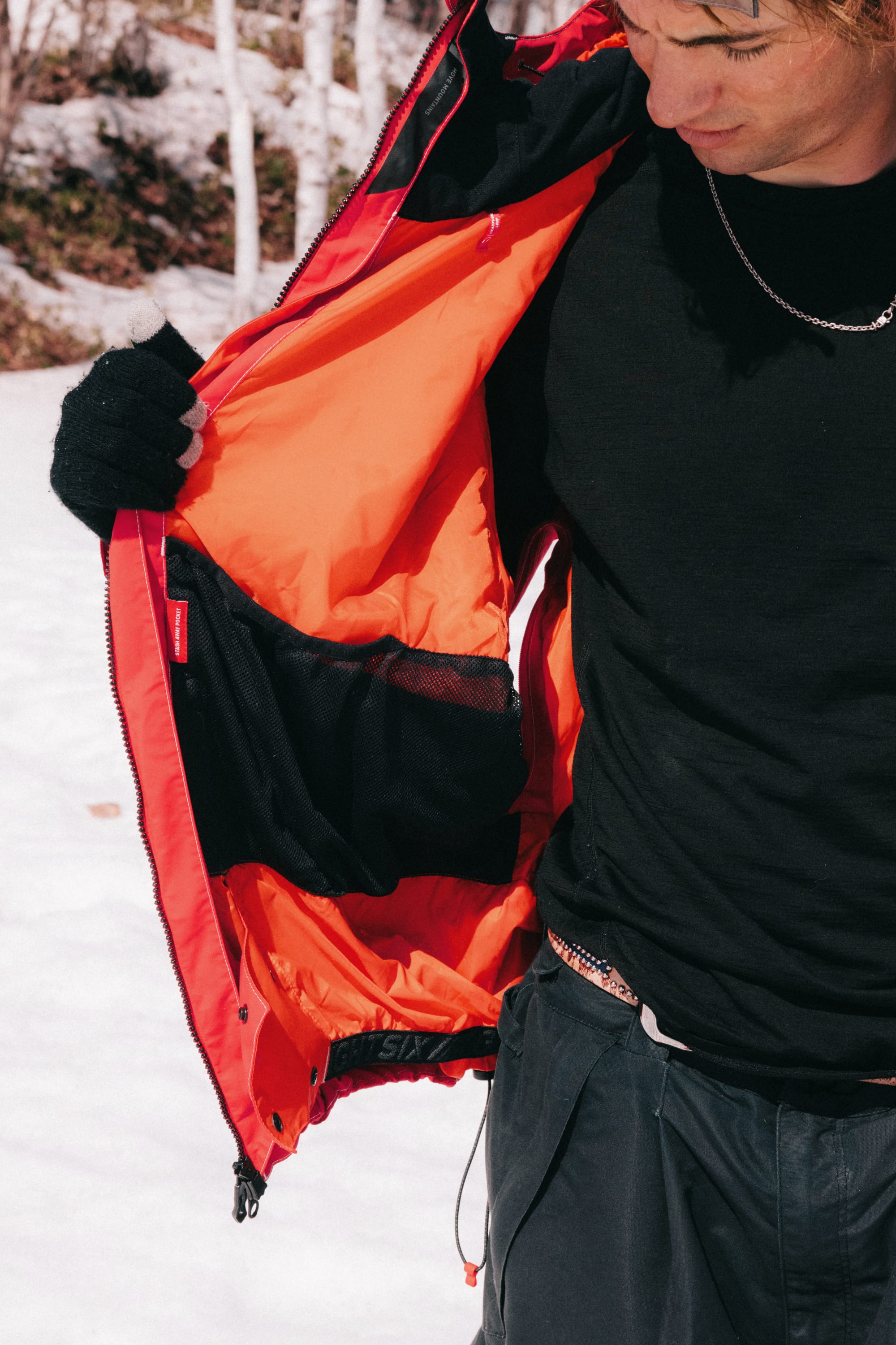 A person in black clothes and gloves holds open a 686 Dojo® GORE-TEX Jacket, revealing its inner mesh pockets, while standing on snow.