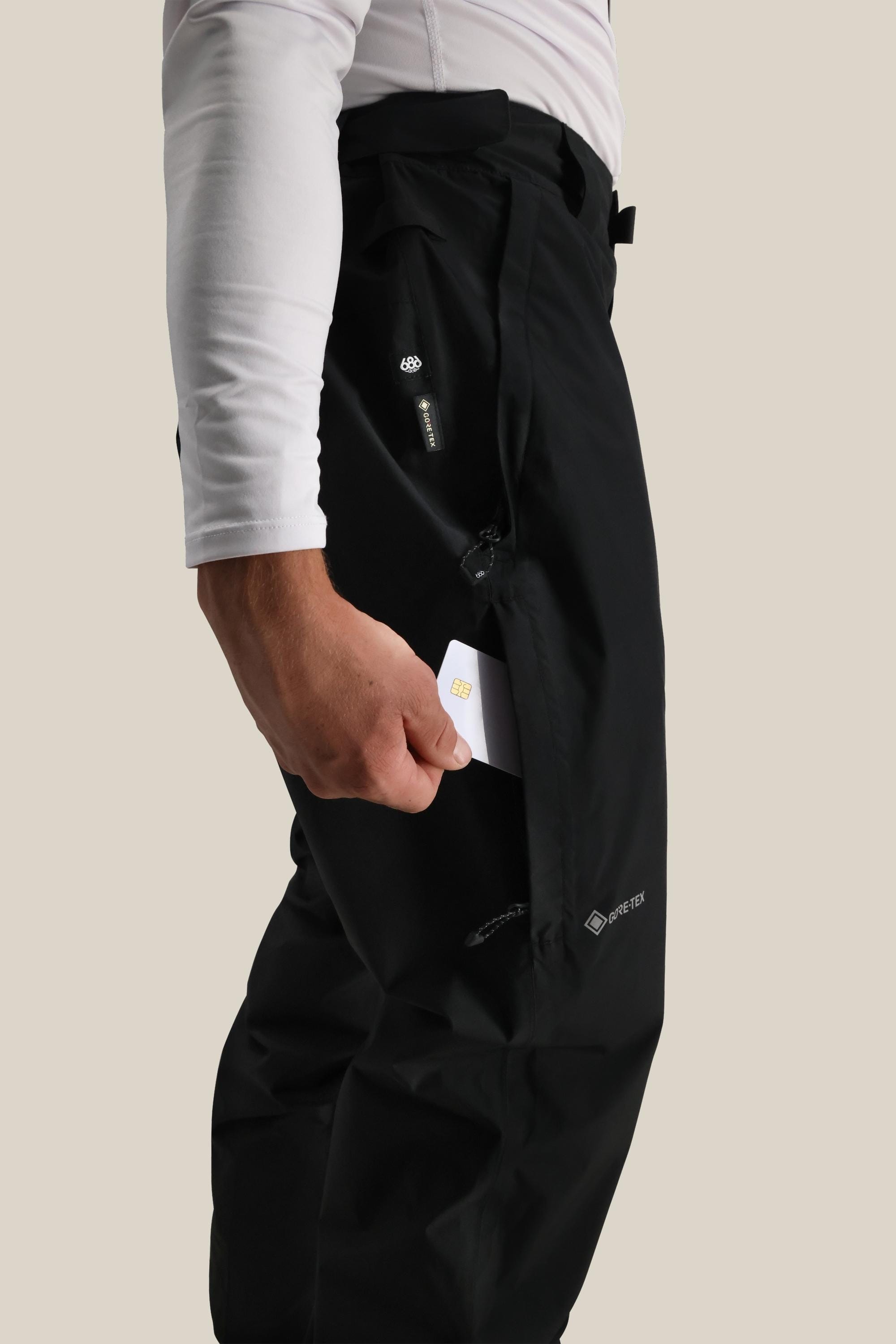 A person wearing 686 Mens GORE-TEX Core Shell Pant in black and a long-sleeve white shirt slips a small white card into the zippered side pocket. The background is plain beige.