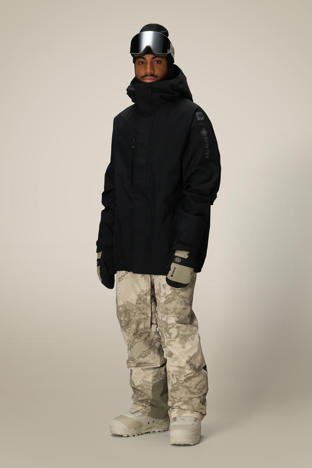A person stands against a plain background wearing a black hooded jacket, 686 Mens GORE-TEX Core Shell Pant in tan and gray camo, light gray gloves, beige snow boots, and black ski goggles on their forehead.